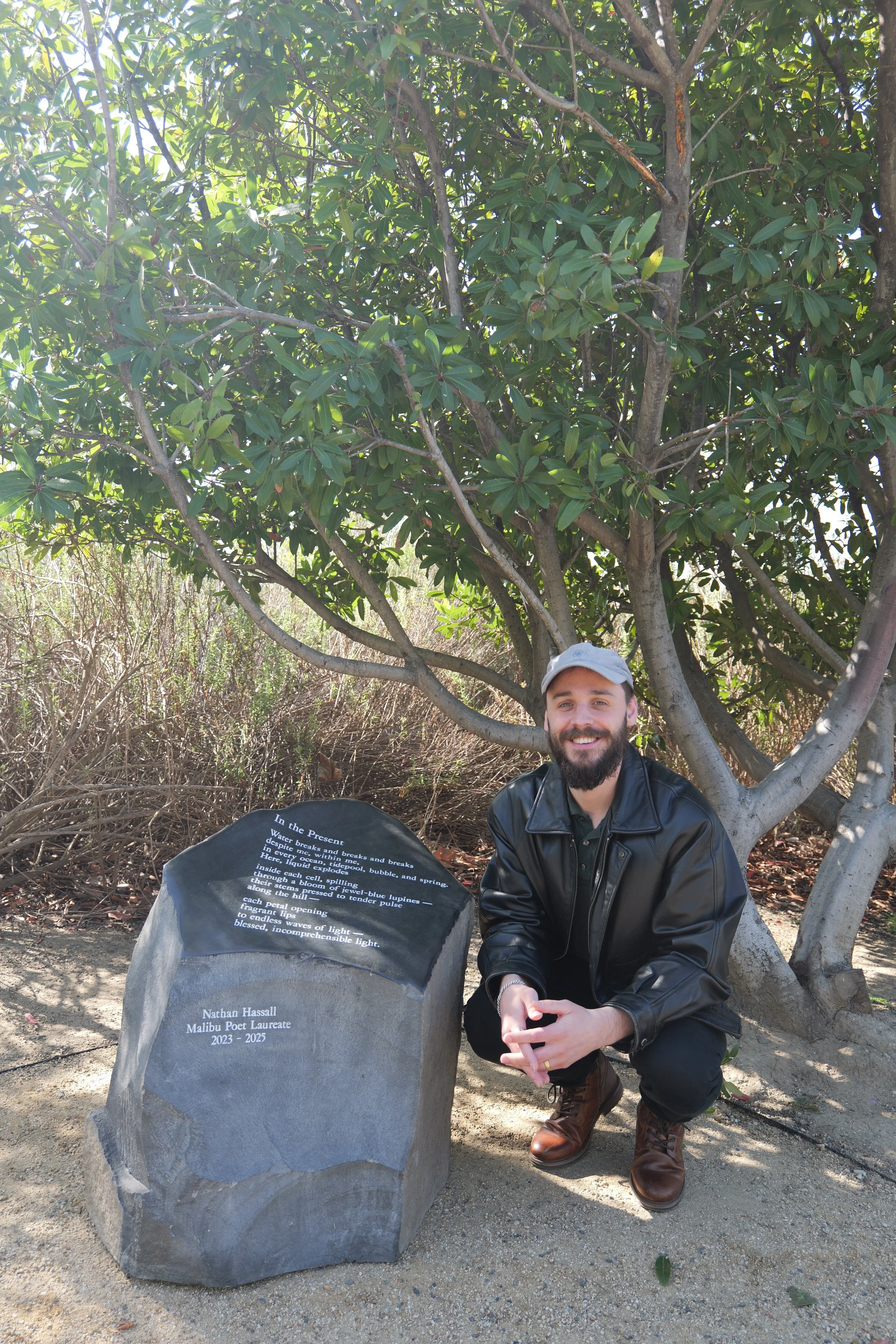 Malibu Poet Laureate Emeritus and Founder of The Poetry Vessel, Nathan Hassall, wearing a gray cap and black leather jacket, squatting beside his poetry stone outdoors under a tree with green leaves.