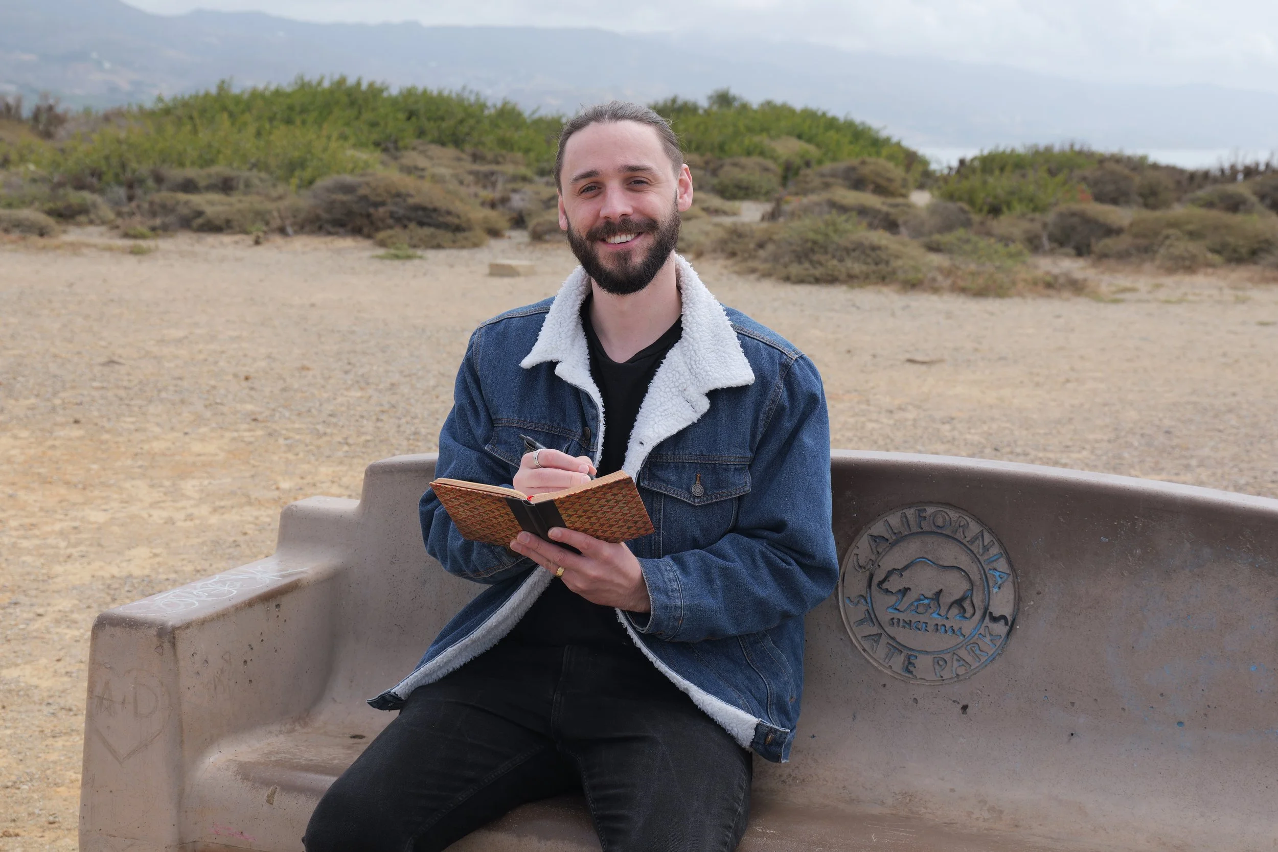 Malibu Poet Laureate Emeritus and Founder of The Poetry Vessel, Nathan Hassall, wearing a denim jacket with a white fleece lining, sitting on a bench at a park, smiling and holding a small notebook and pen.
