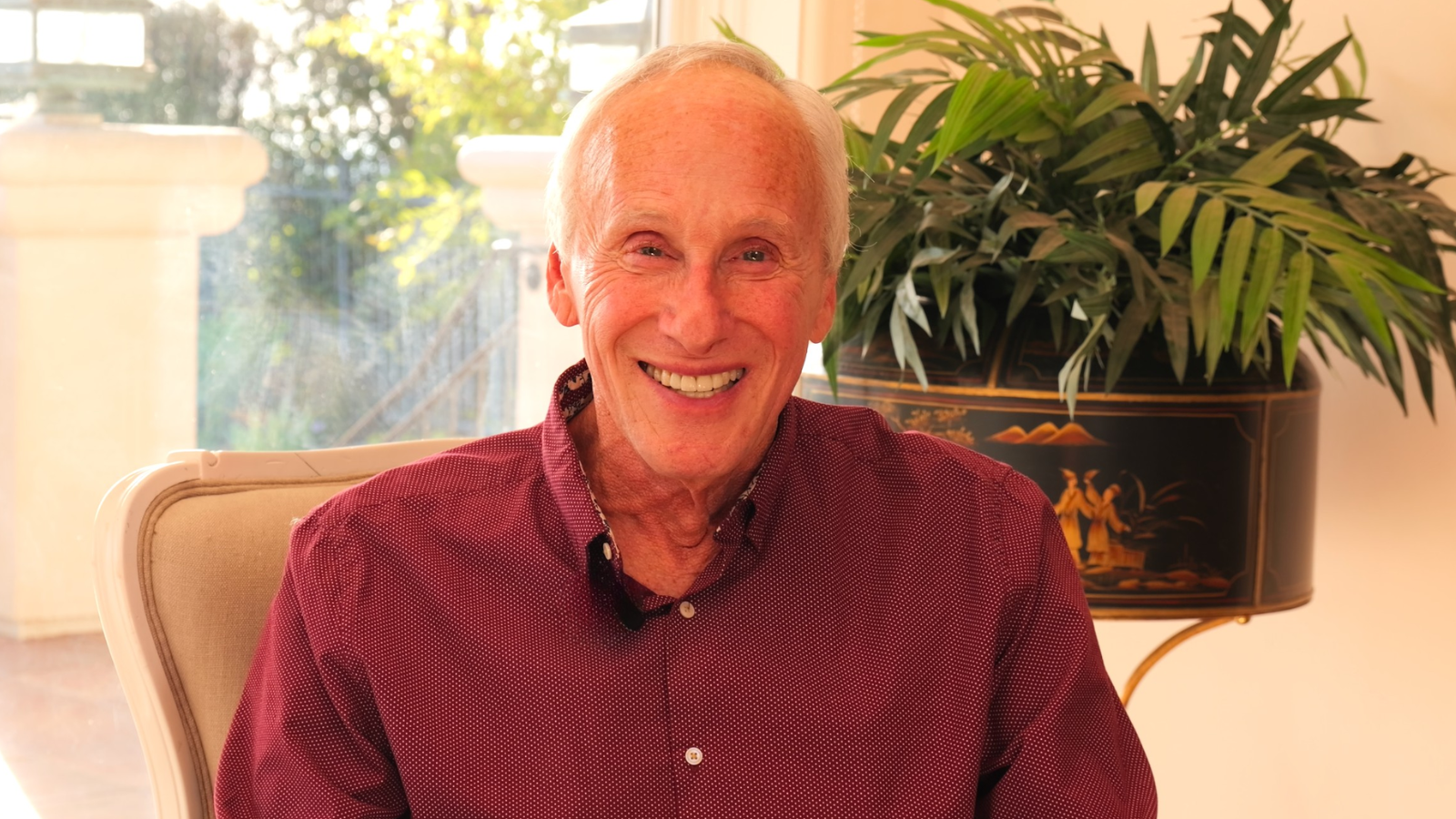 Mark Waldman, national best-selling author and neuroscience researcher sitting in a chair in front of a window with sunlight, and a large potted plant with green leaves behind him.