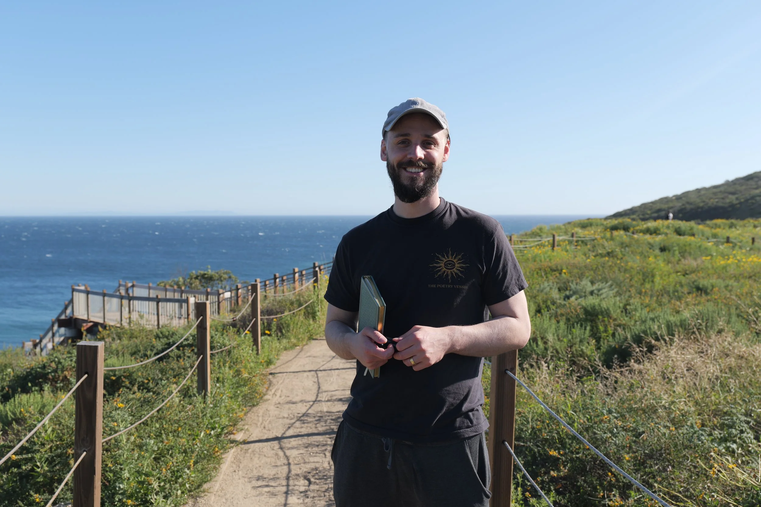 Nathan Hassall, Malibu Poet Laureate Emeritus and Founder of The Poetry Vessel, holding a poetry notebook, standing on a pathway with a wooden fence, overlooking the ocean and a green hillside with yellow flowers.