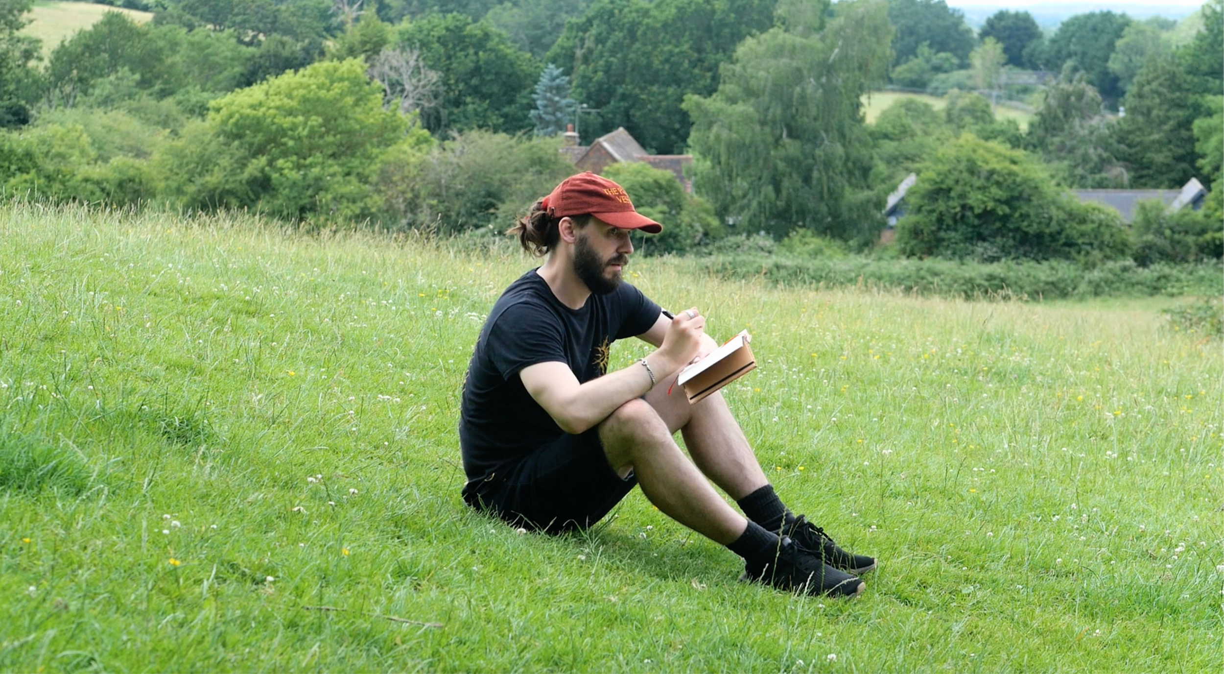 Malibu Poet Laureate Emeritus and Founder of The Poetry Vessel, Nathan Hassall, sitting on a grassy field writing in a notebook with trees, houses, and hills in the background.