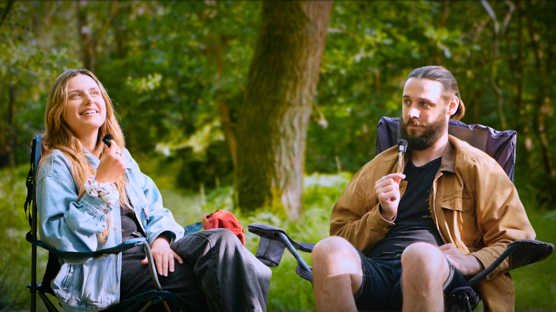 Rachael and Nathan Hassall, Malibu Poet Laureate Emeritus, sitting outdoors in St. Leonard's forest, both holding podcast microphones and smiling, wearing casual clothing and camping chairs nearby.