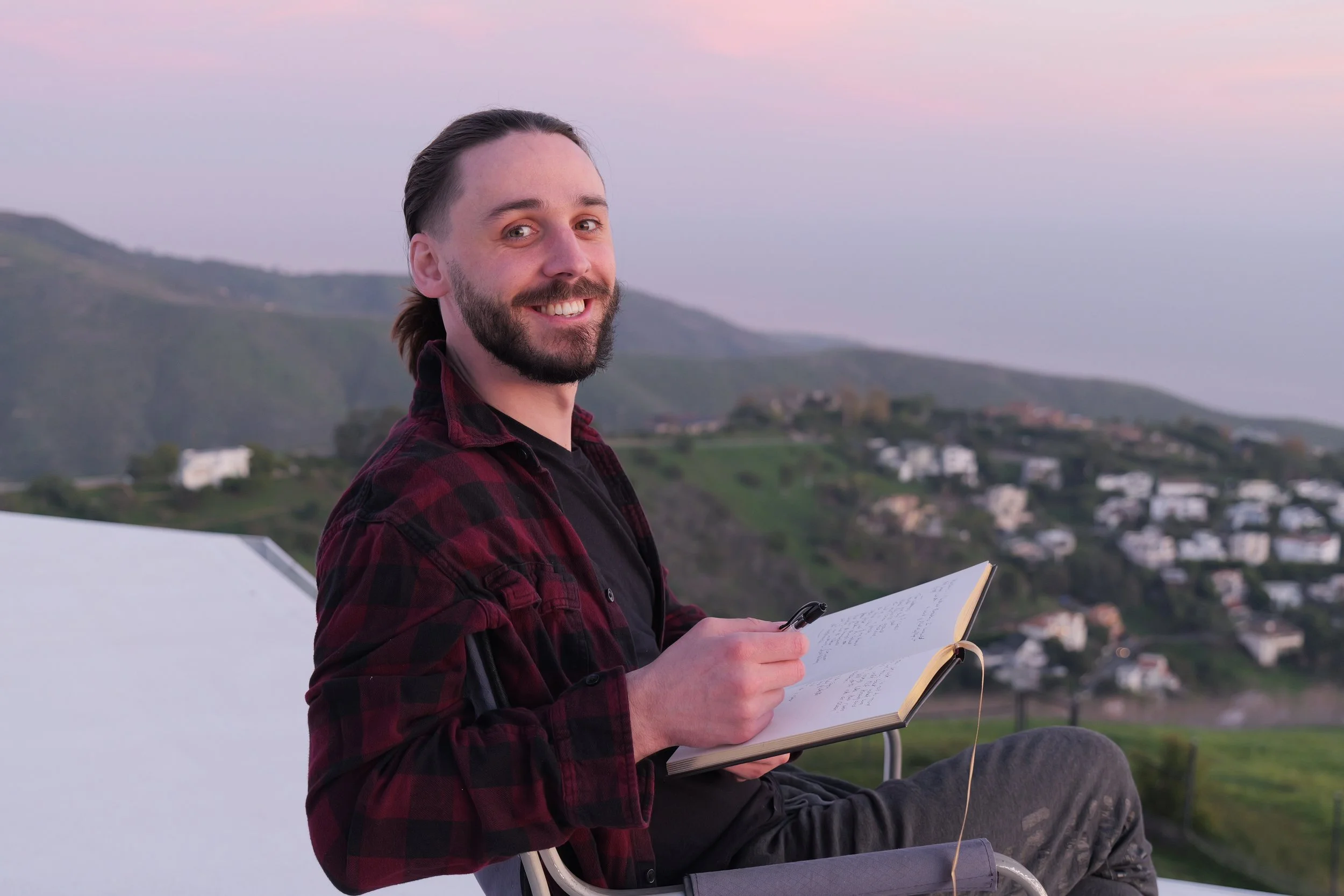 Nathan Hassall, Malibu Poet Laureate Emeritus and Founder of The Poetry Vessel, sitting outdoors at sunset, holding an open notebook and a pen, with scenic hills and houses in the background.