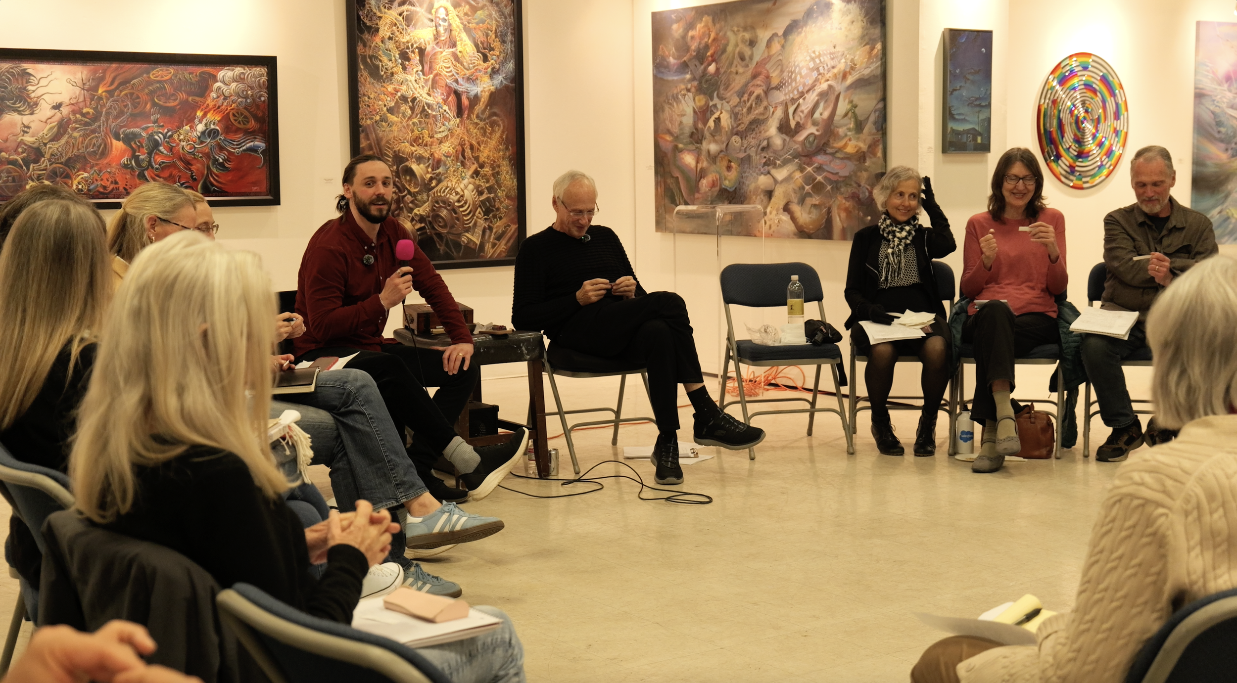 A group of people sitting in a semi-circle in an art gallery, participating in a discussion or presentation, with colorful abstract paintings hanging on the walls behind them.