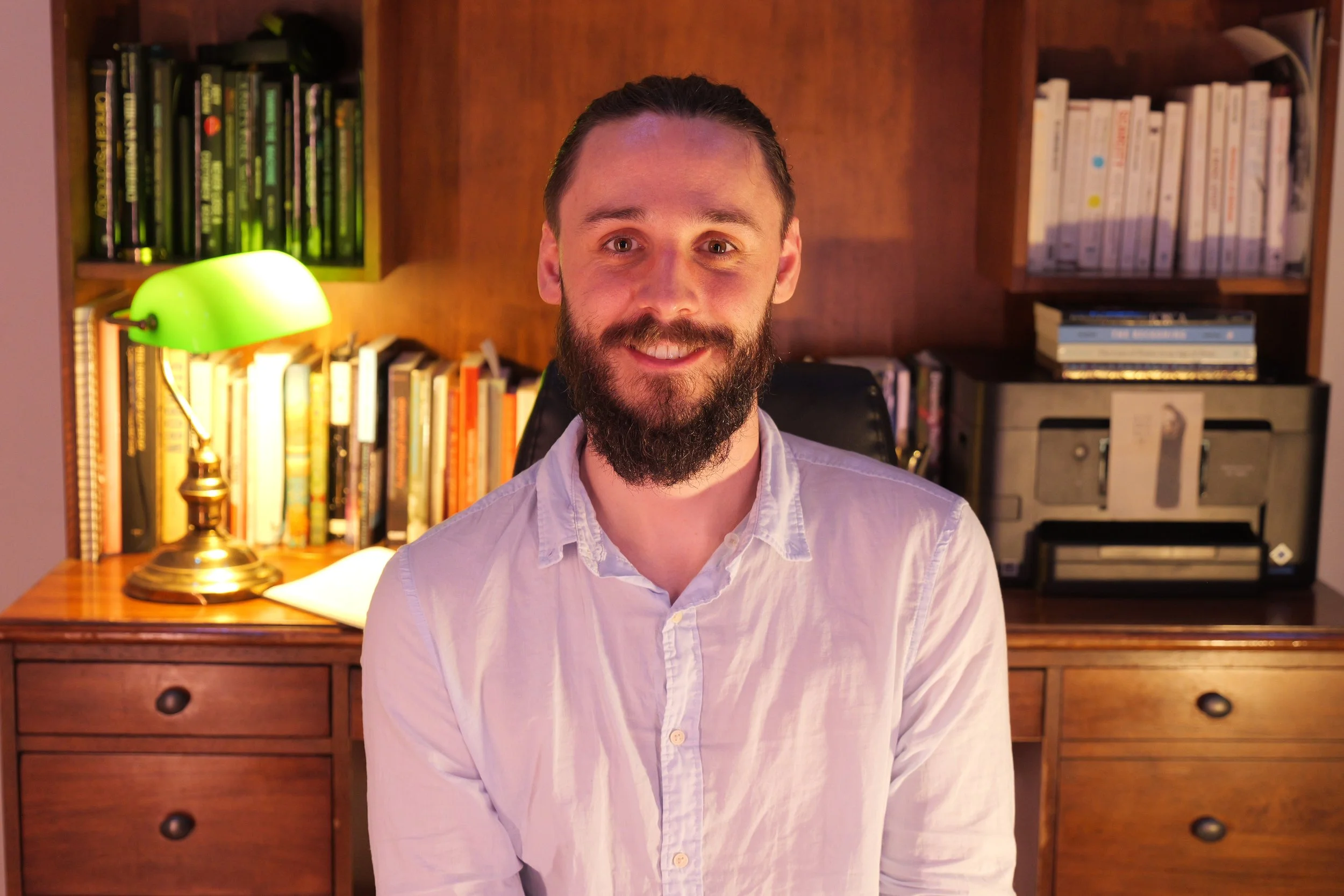 Malibu Poet Laureate Emeritus and Founder of The Poetry Vessel, Nathan Hassall, smiling and sitting in a room with wooden furniture, bookshelves, and a green banklamp.