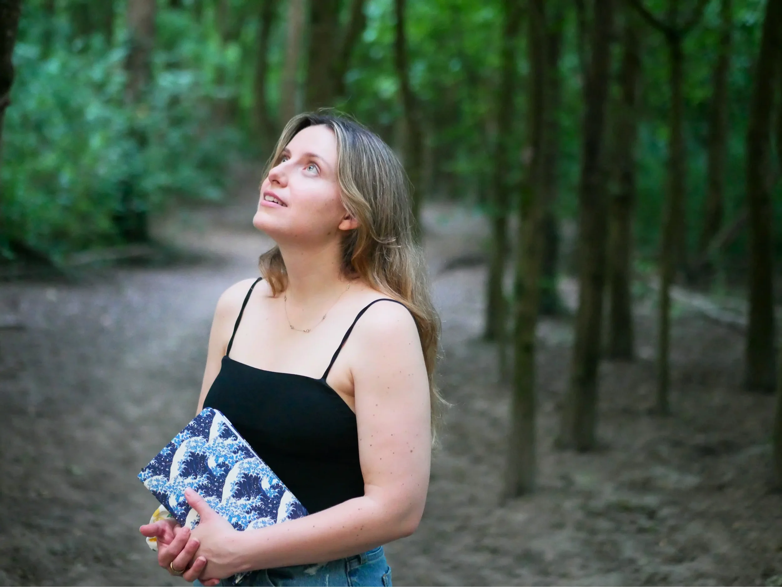 Rachael Hassall, wife of Malibu Poet Laureate Emeritus, Nathan Hassall, in a black spaghetti strap top holding a blue and white patterned book or notebook, standing outdoors in a green forest.