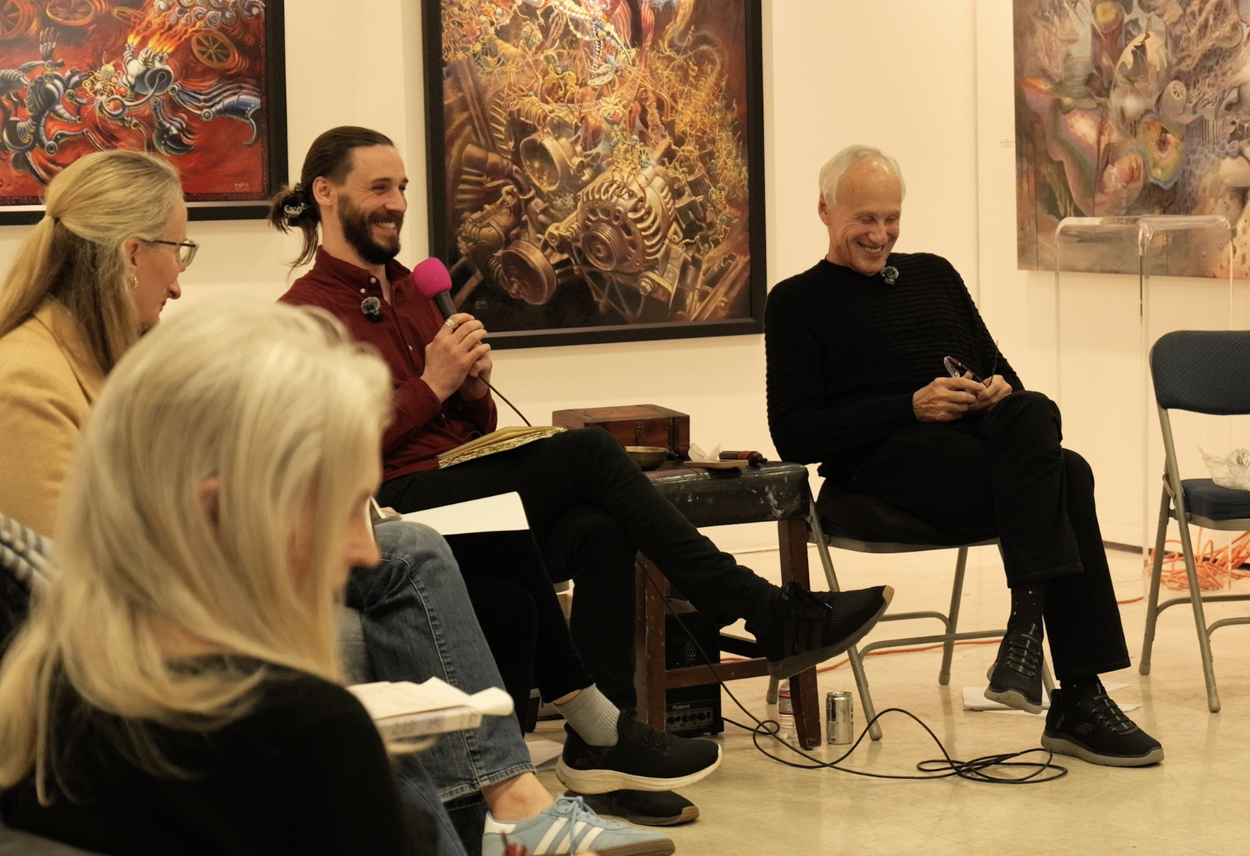 Nathan Hassall, Malibu Poet Laureate Emeritus and Founder of The Poetry Vessel leading a poetry writing workshop in an art gallery or museum, engaging in a discussion or presentation, with colorful abstract paintings hanging on the wall behind them