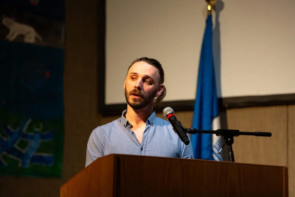 Nathan Hassall, Malibu Poet Laureate Emeritus and Founder of The Poetry Vessel, speaking at a podium with a microphone in a large auditorium.