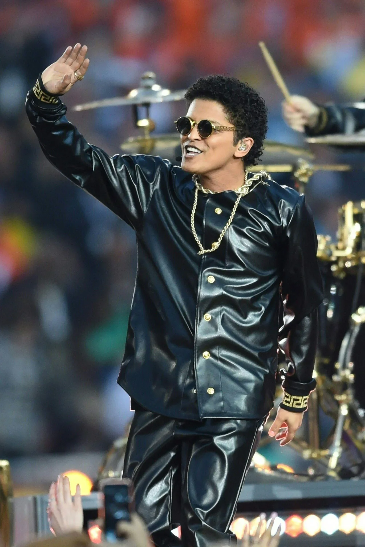 A male performer with curly hair, wearing sunglasses, a black leather shirt and pants, gold jewelry, waving to fans at a concert.