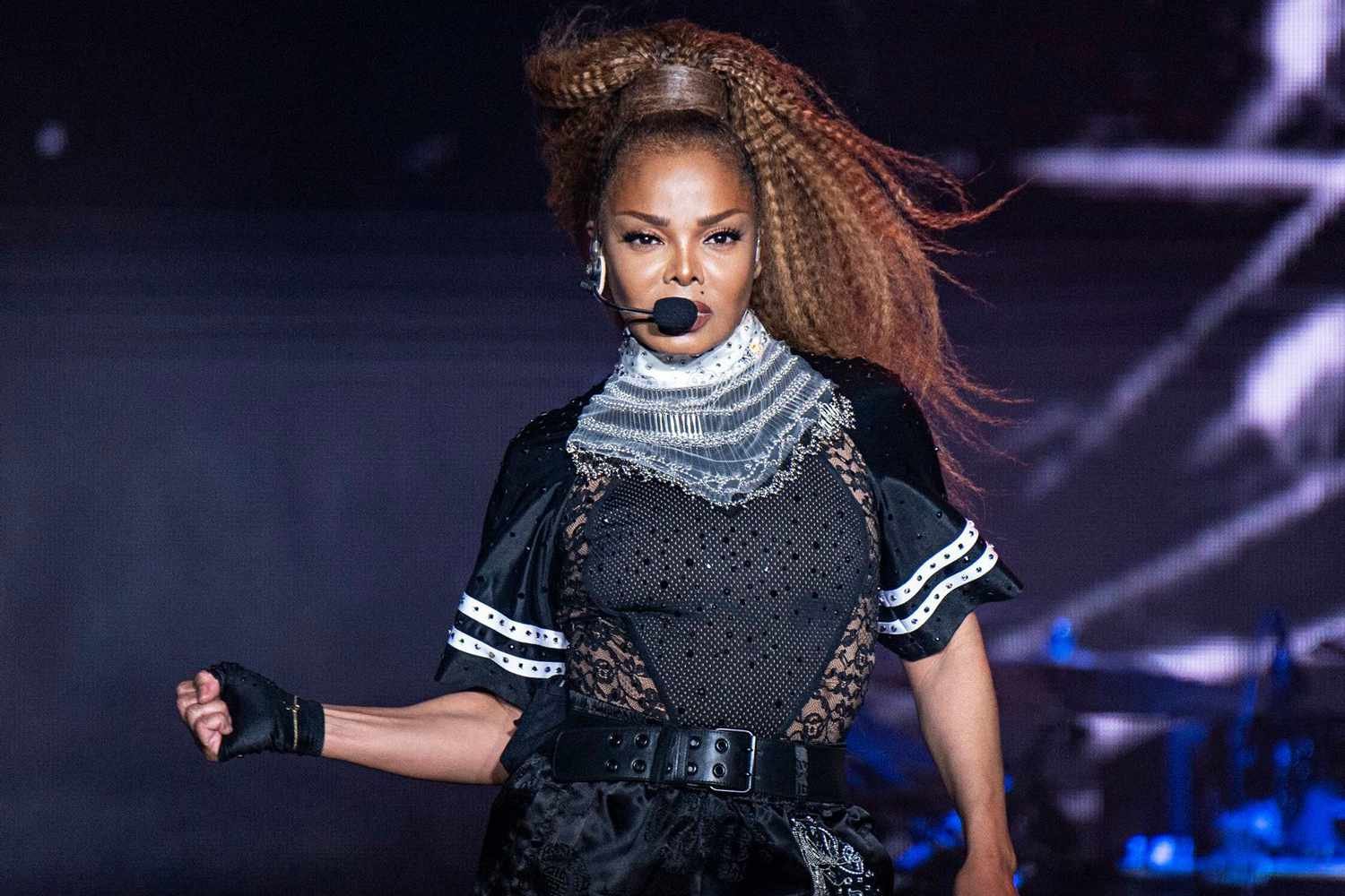 A woman with long, braided hair performing on stage, wearing a black and lace outfit with a silver scarf around her neck, holding a microphone headset, with stage lights in the background.