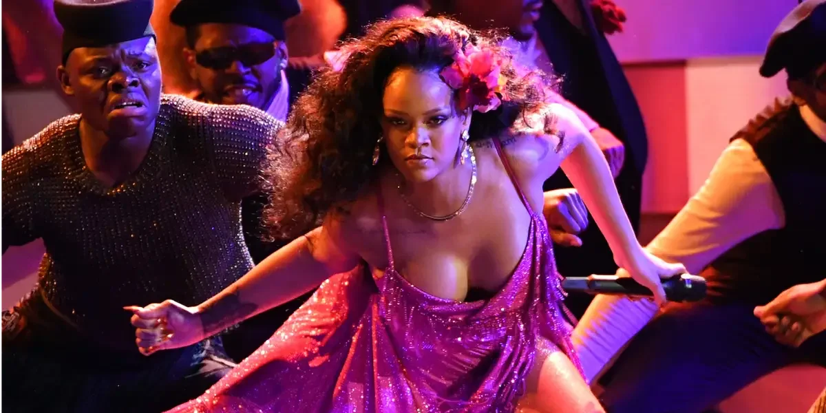A woman in a sparkly purple dress holds a microphone and performs on stage, surrounded by a crowd at a lively event.