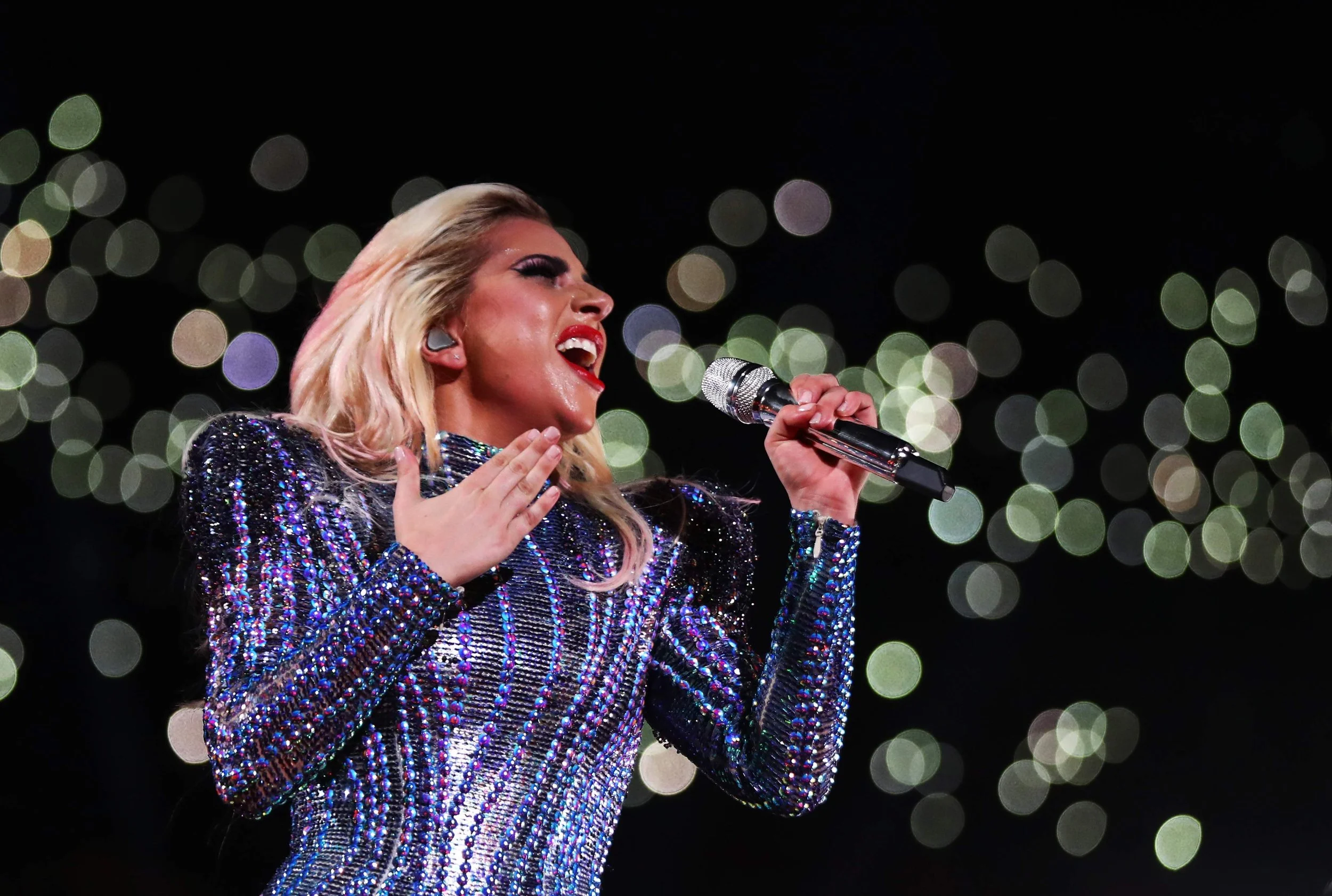 A female singer performing on stage, holding a microphone with her right hand, dressed in a dark, shiny, and colorful beaded outfit, with bokeh lights in the background.