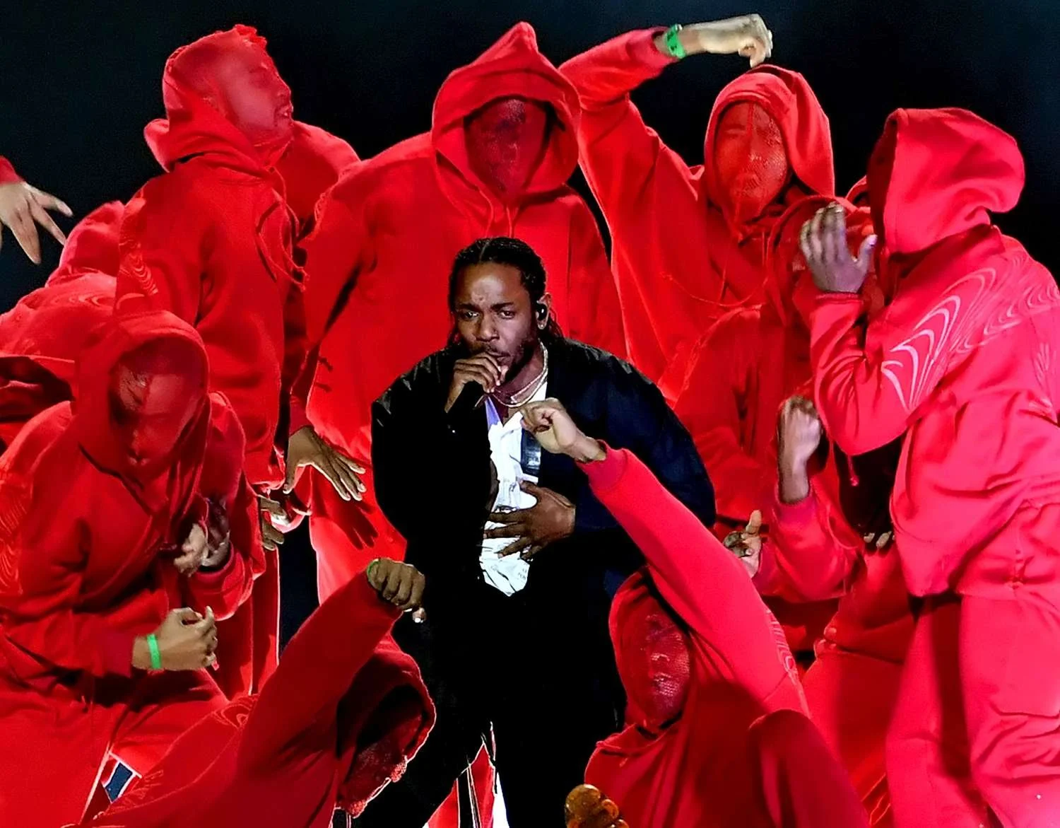 A male performer wearing a black jacket and white shirt, holding a microphone, surrounded by individuals in red hoodies and pants, some with their faces covered, on a dark stage.