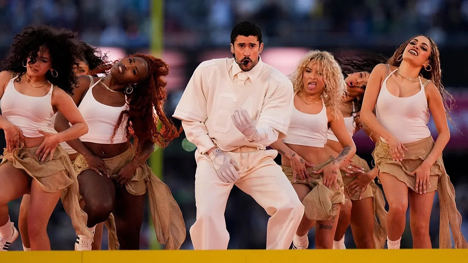 Man wearing white dance outfit and gloves performing with a group of five women dancer in white crop tops and tan skirts, on a stage or set with a blurred background.