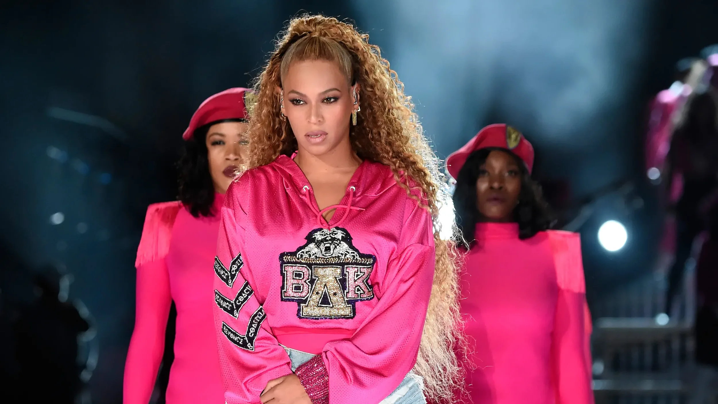 A woman with curly hair in a pink sports jersey with embroidered patches and Greek letters, standing with arms crossed, flanked by women in pink uniforms and hats, in a photo with dramatic lighting.