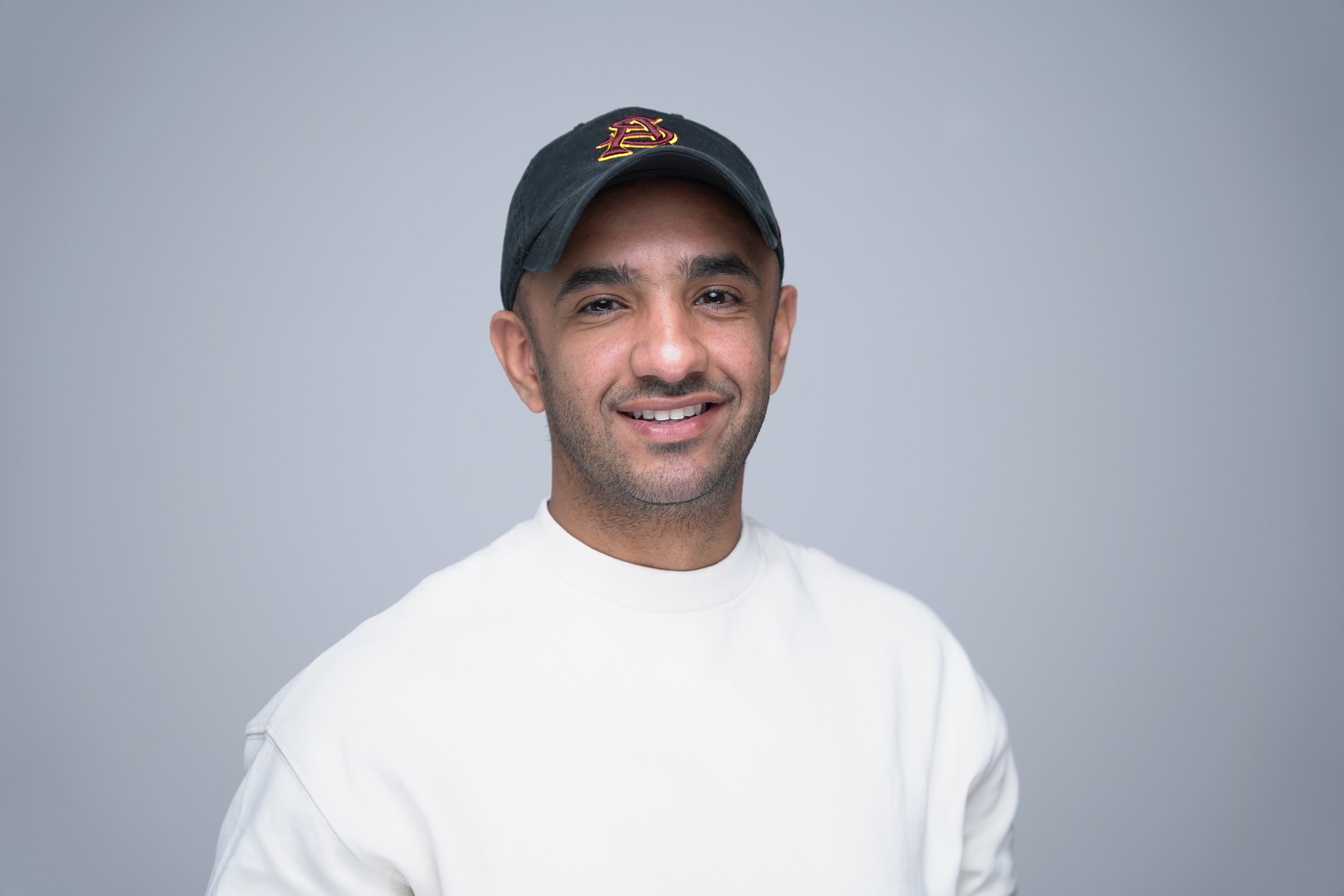 A smiling man wearing a black baseball cap with a red and yellow emblem, dressed in a white sweatshirt, standing against a plain gray background.