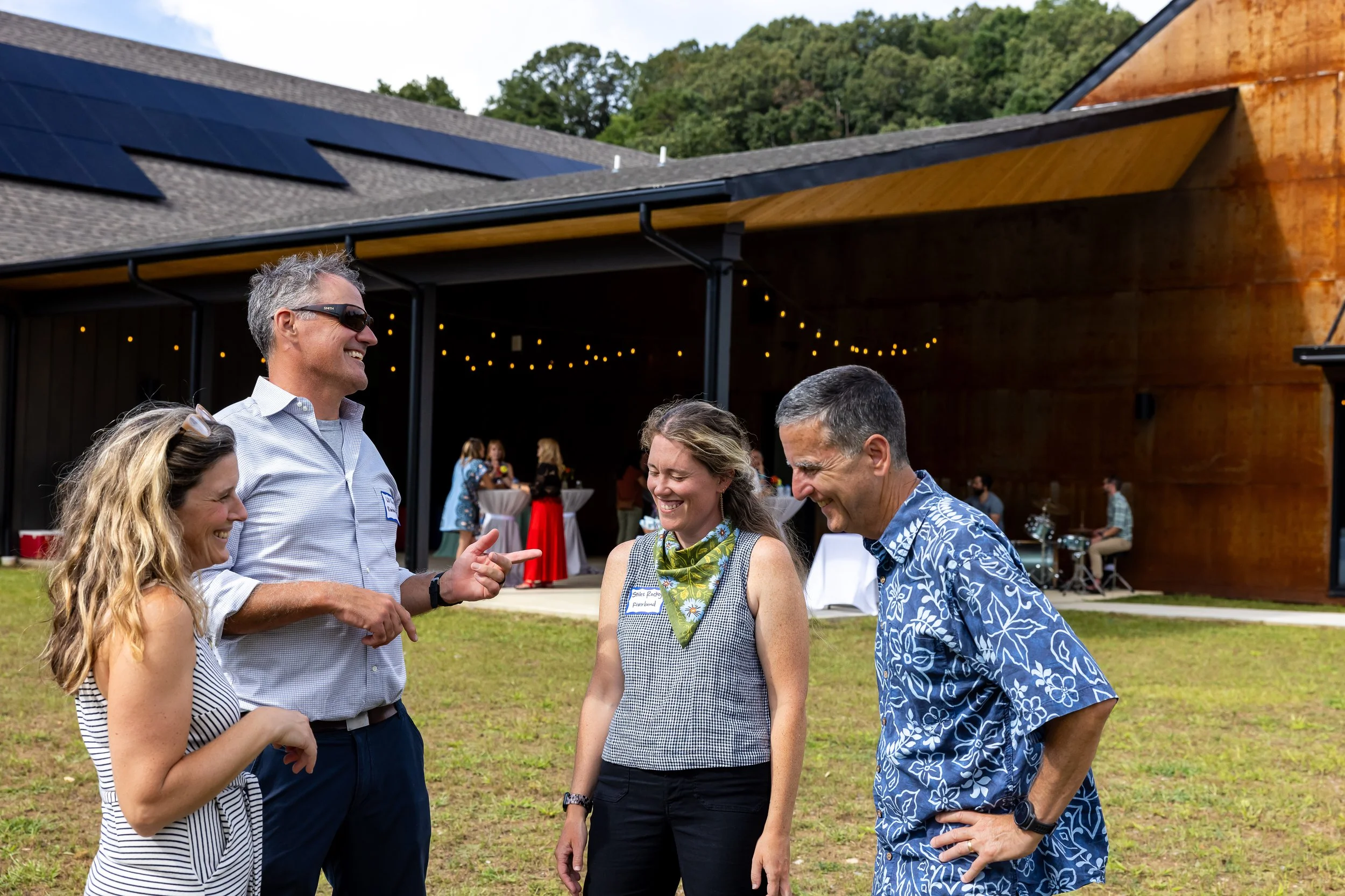 A group of five people outdoors at a social gathering, smiling and talking, with a building and trees in the background.