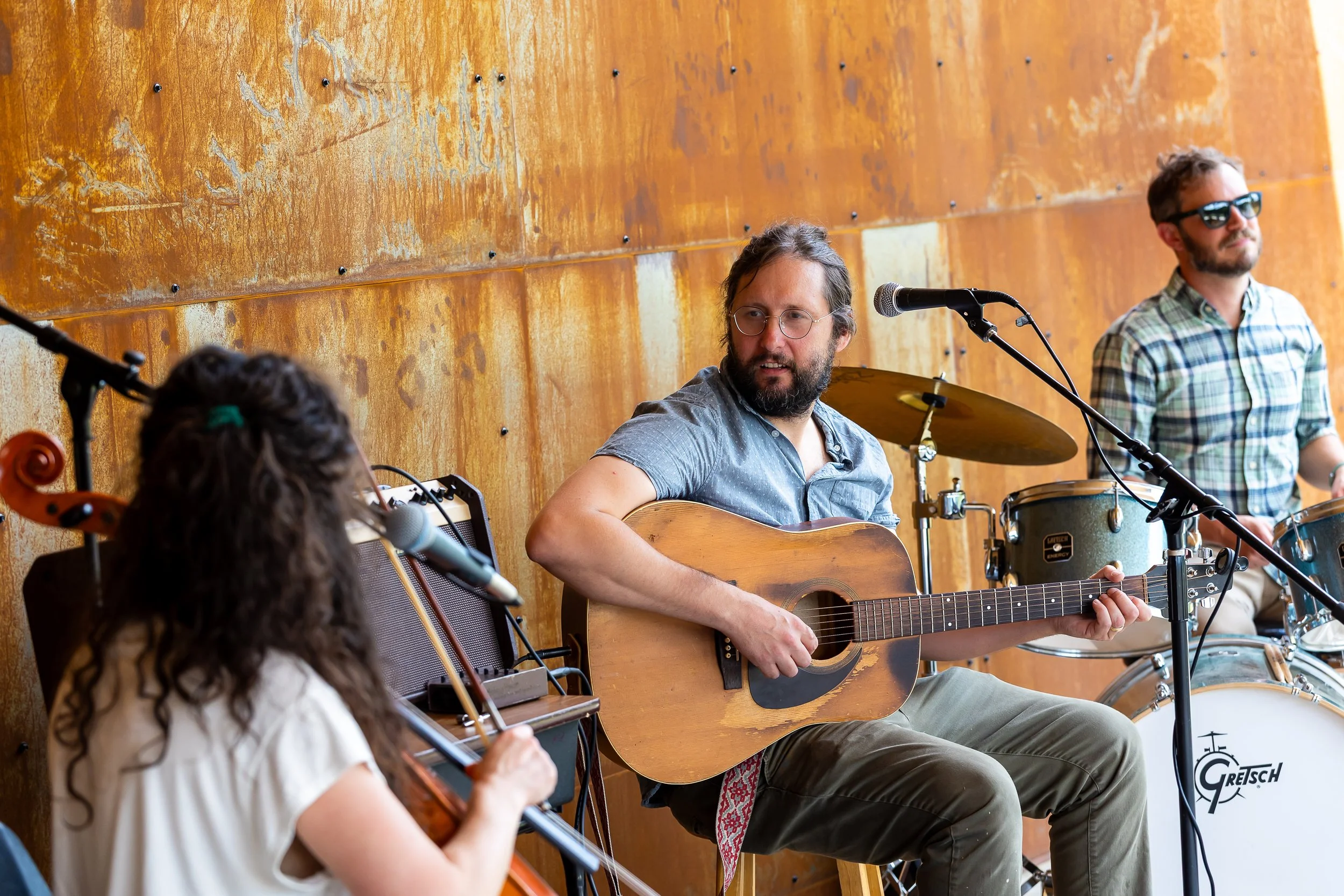 Three musicians performing on stage, two men with guitars and a woman with a violin, in front of a rusty metal wall.