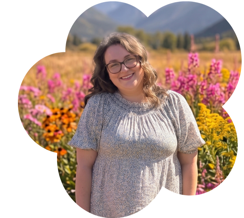 A woman with glasses and wavy brown hair smiling in a field of colorful flowers with mountains in the background during daytime.
