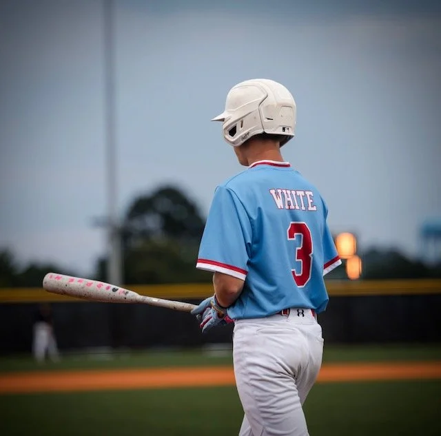 A young baseball player wearing a white Rawlings helmet and a blue jersey with 'WHITE' and the number 3 on the back, holding a Combat baseball bat on a baseball field during dusk.
