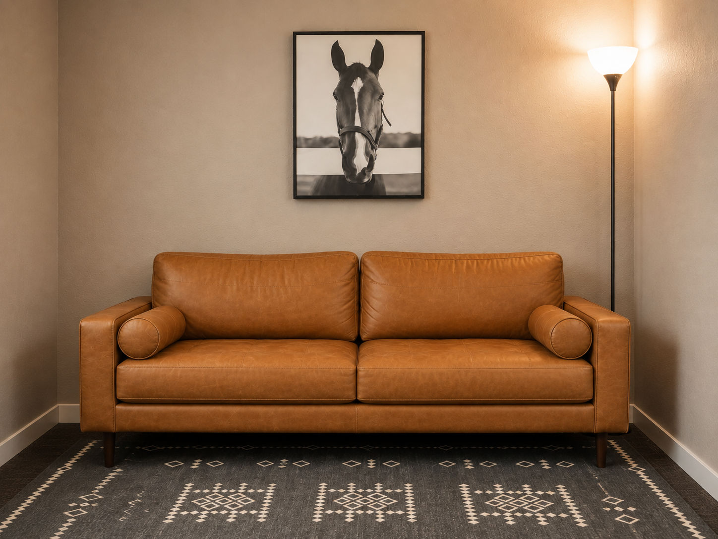 A living room scene with a tan leather sofa, two round bolster pillows, a black and white framed photograph of a horse, a tall floor lamp with a white shade, and a patterned rug on the floor.