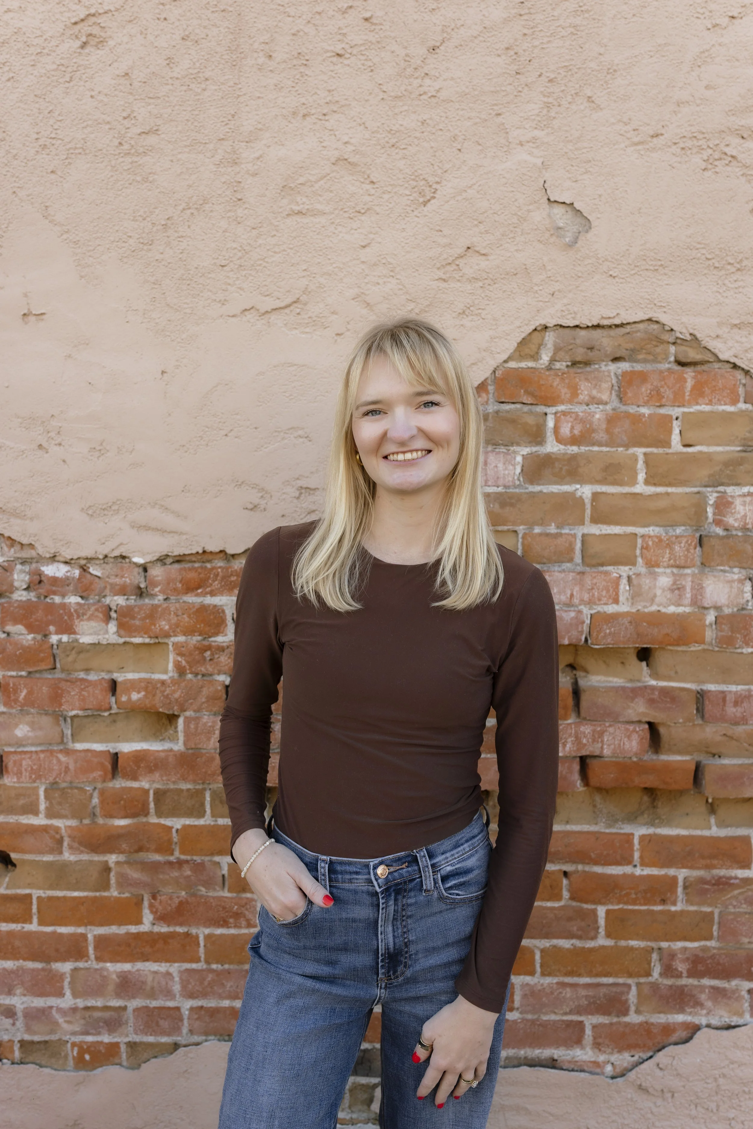Smiling blonde woman in a brown top and jeans standing against a weathered brick wall
