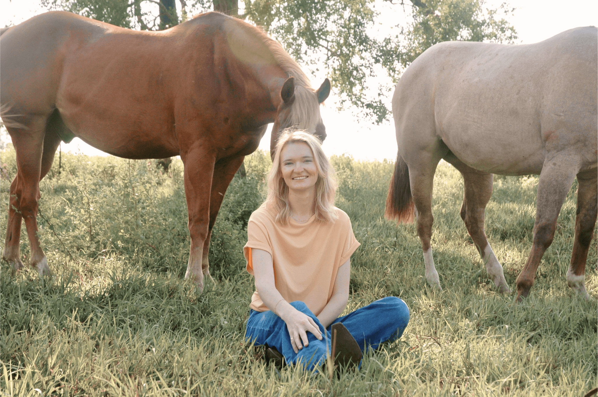 Hallie Mesersmith, LCSW, LMHP sitting on green grass outdoors, smiling, with two horses standing behind her, one brown and white and the other gray.