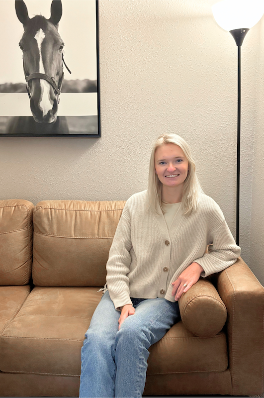 Hallie Mesersmith, LCSW, LMHP sitting on a tan leather couch in a living room, smiling at the camera. Behind her is a framed black and white photograph of a horse's head. To her right is a tall floor lamp.