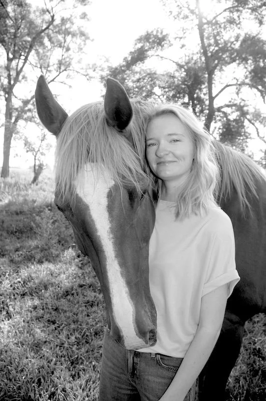 Hallie Mesersmith, LCSW, LMHP standing next to a horse on a grassy field with trees in the background.