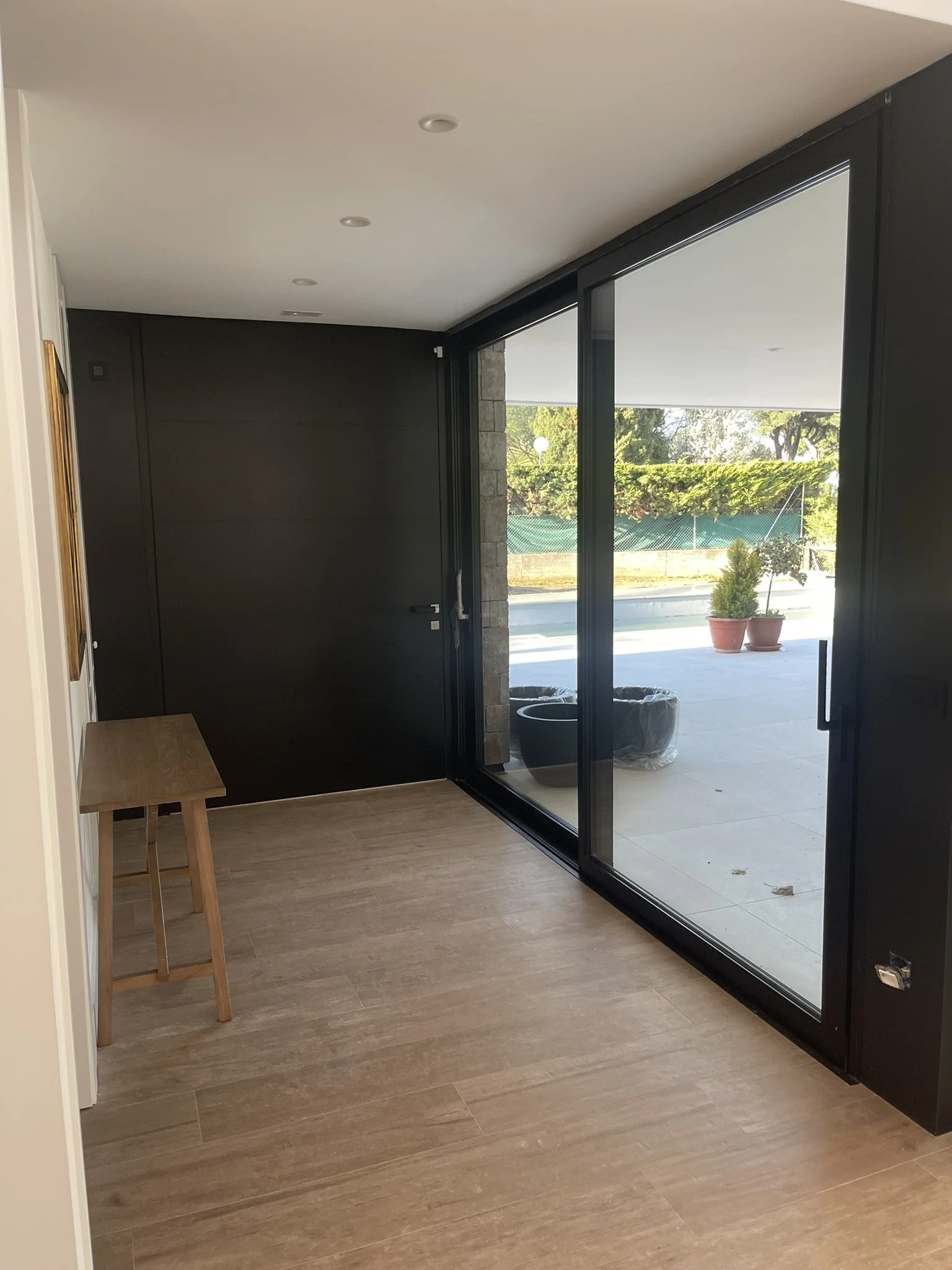 Interiores de una casa con piso de madera clara, pared gris, y gran ventanal con vista a un jardín con árboles y zacate. En la esquina izquierda, un pequeño banco de madera y una pintura en la pared.