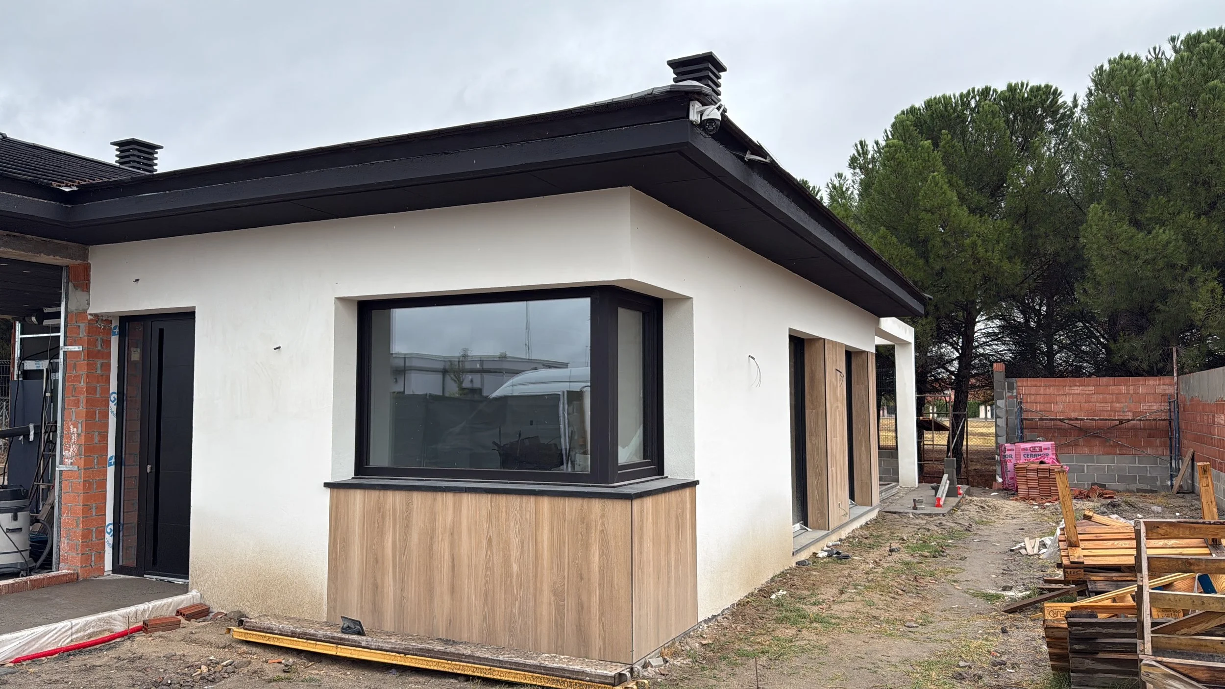 Casa en construcción con paredes blancas, ventanas grandes, y detalles en madera, en un área con árboles y materiales de construcción alrededor.