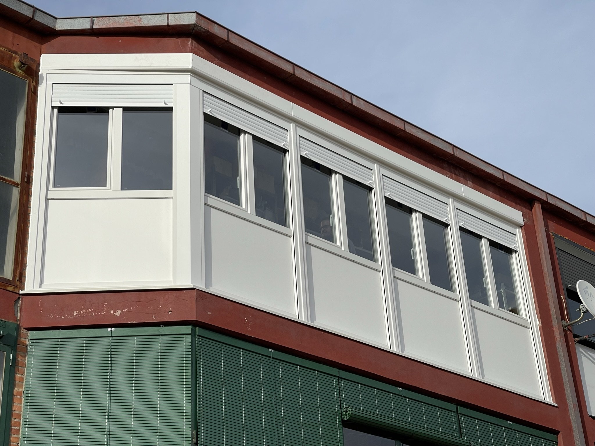 Parte superior de un edificio de varios pisos con ventanas, con paredes en colores rojo, blanco y verde, y un cielo azul en el fondo.