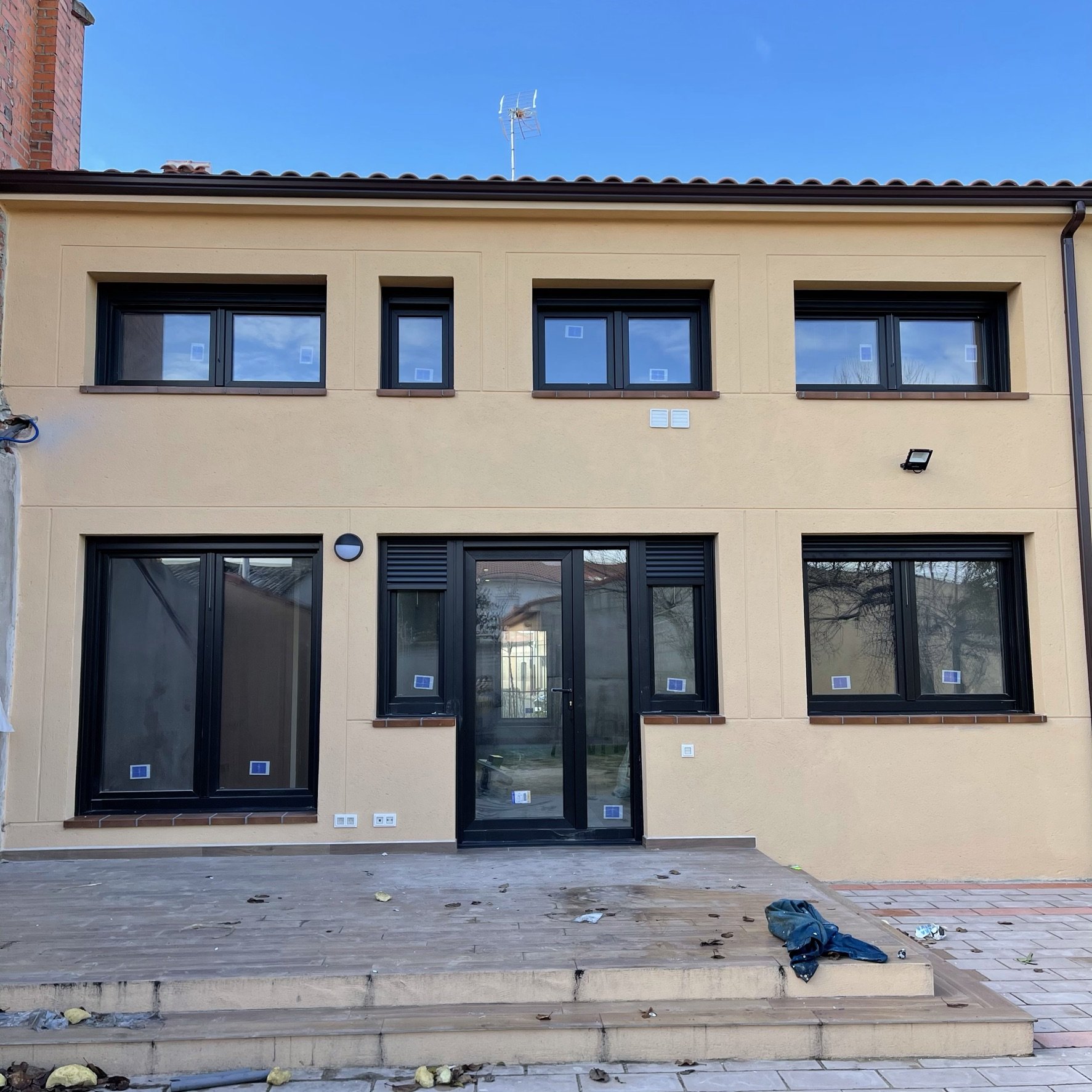 Fachada de una casa con ventanas y puerta de vidrio con marco negro, en construcción o reparación, con basura y herramientas en el suelo