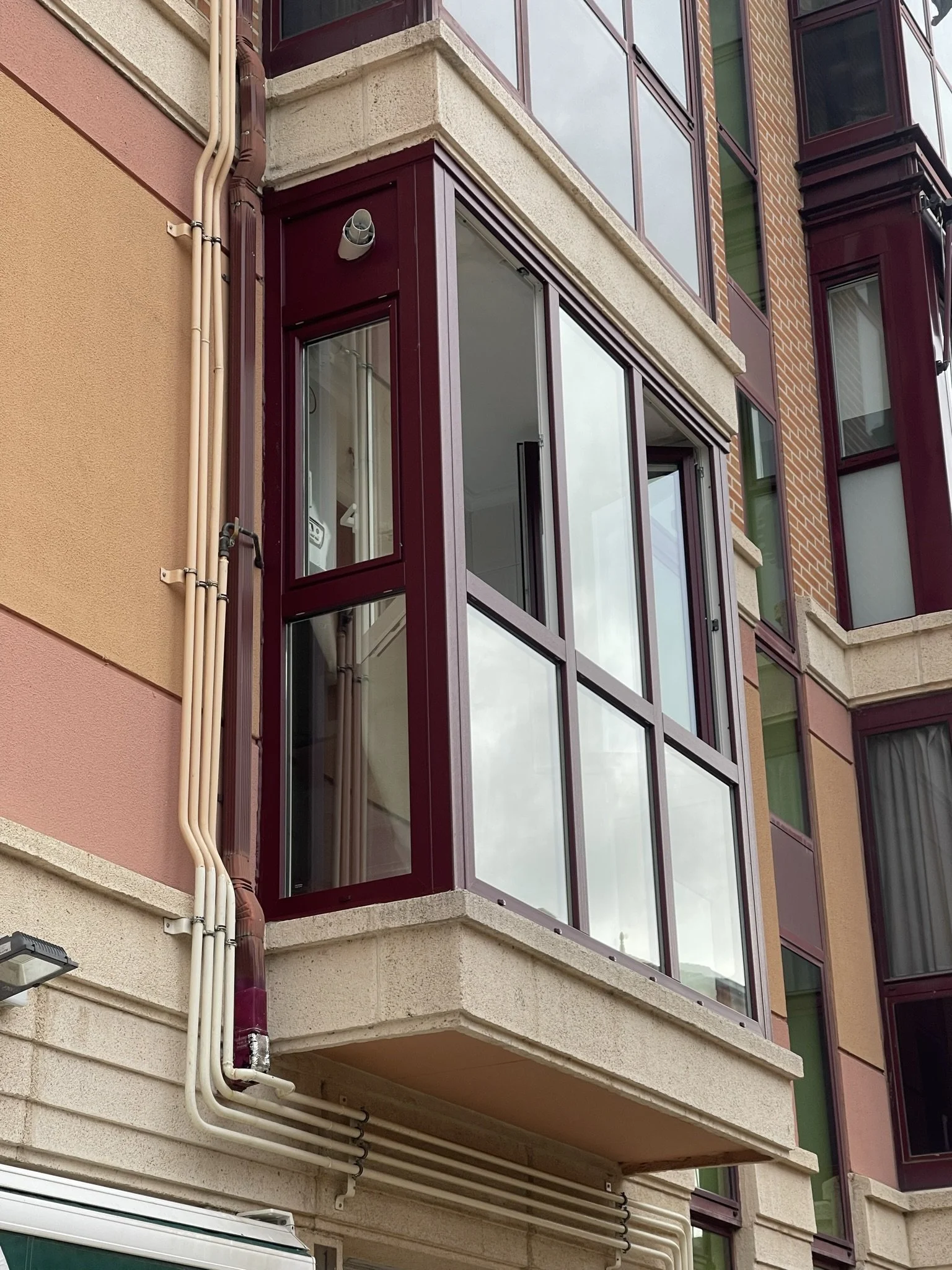 Edificio de varios pisos con ventanas de marco color burdeos y ventanas de vidrio, algunas abiertas, con tuberías y una luz externa en la fachada.