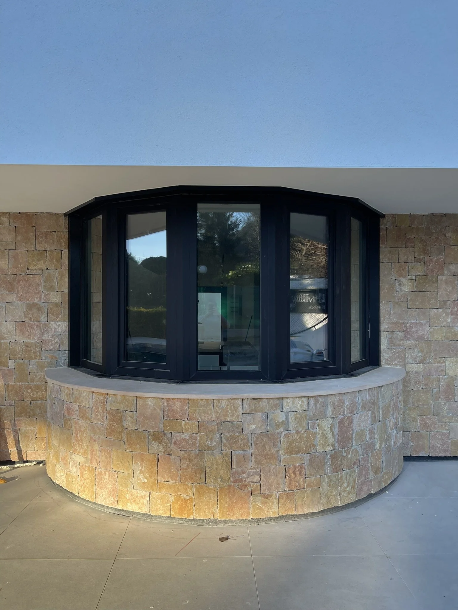 Ventana curva con marco negro en pared de piedra y techo de color beige, vista hacia el exterior con árboles y cielo visible.