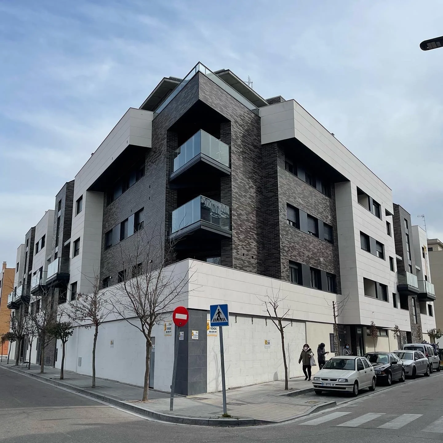 Edificio moderno de varios pisos en una calle urbana, con árboles en la acera y señal de tránsito, y algunas personas caminando.