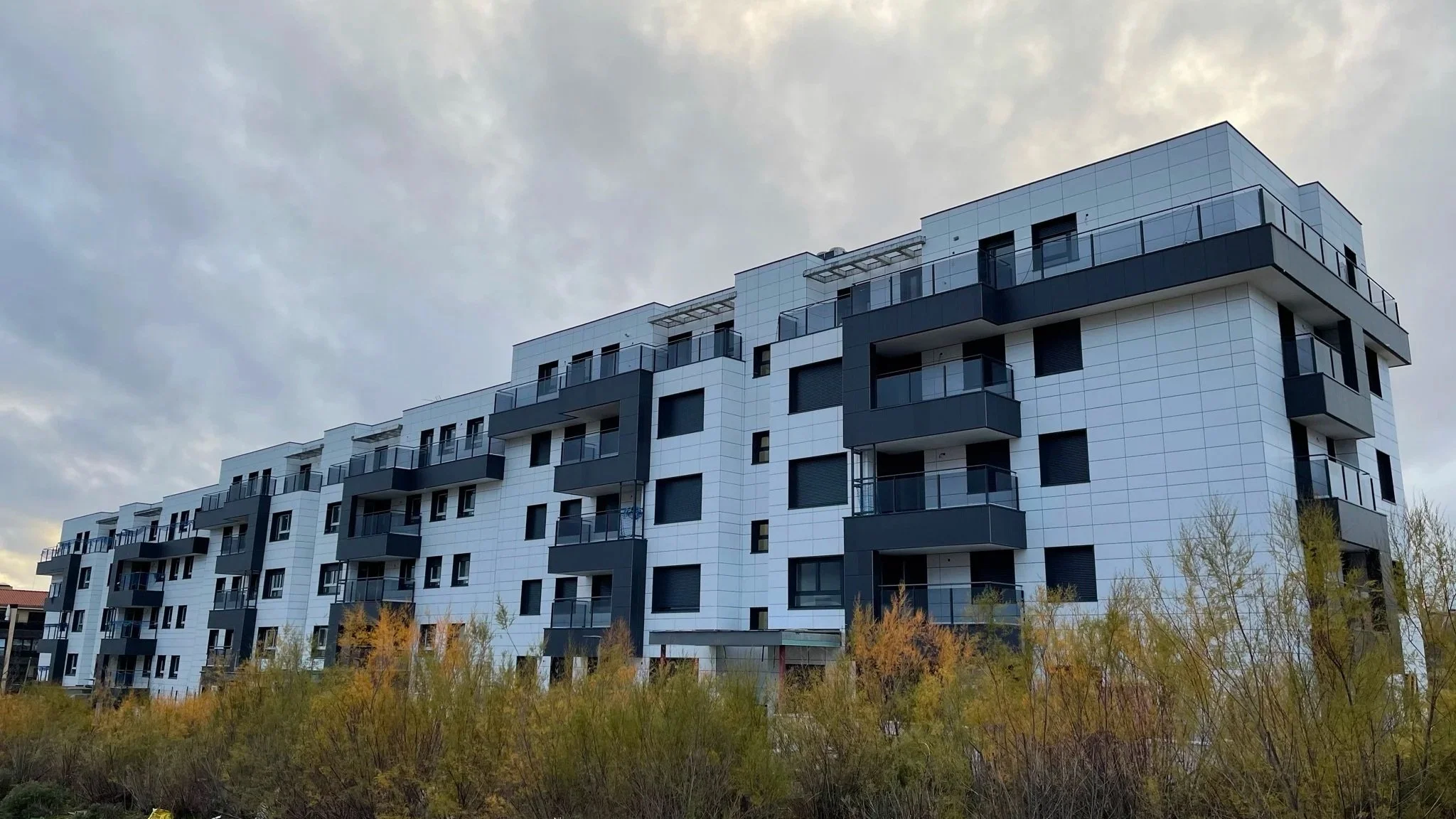 Edificio moderno de apartamentos con balcones y ventanas de vidrio, con árboles en primer plano y cielo nublado de fondo.