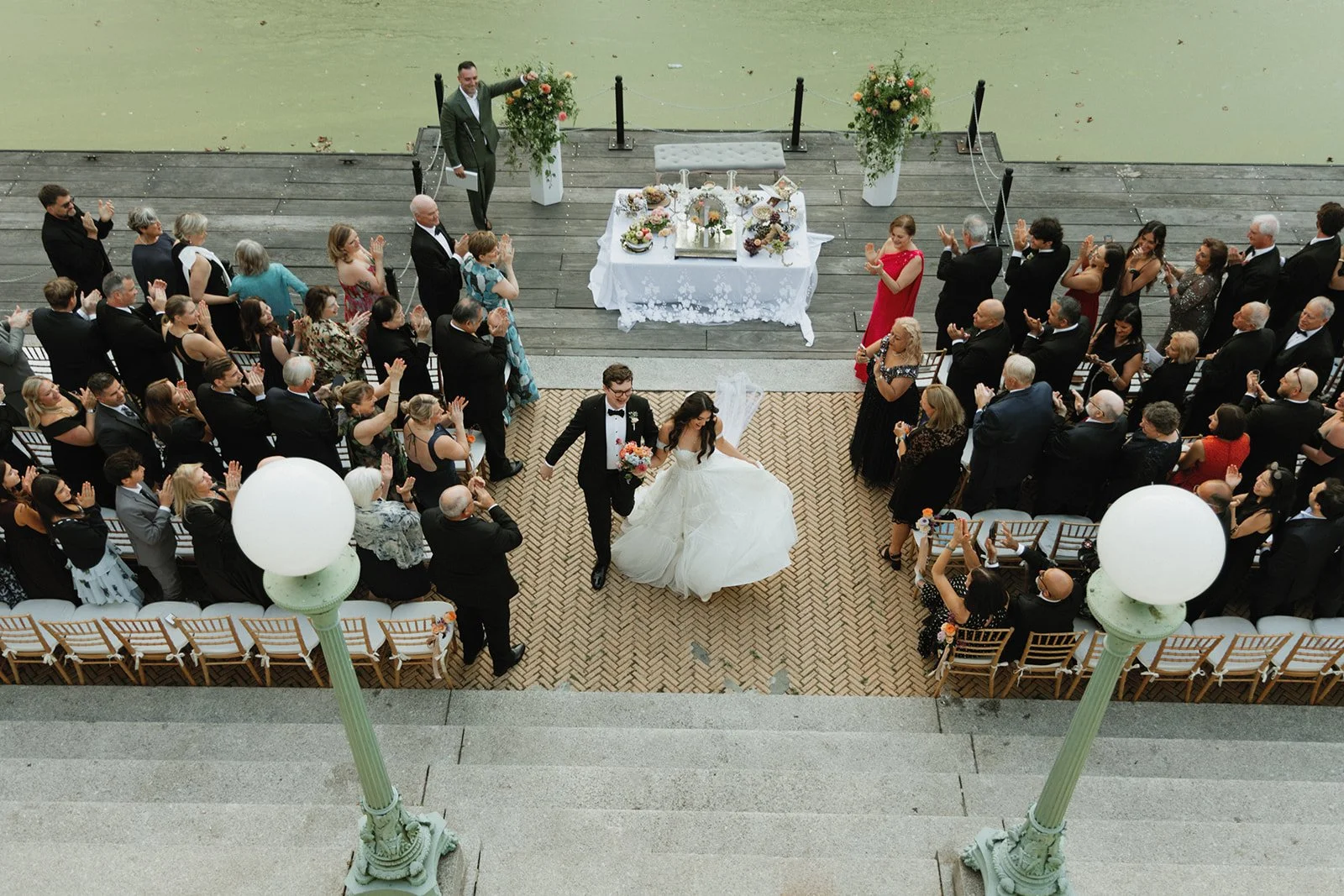 Aerial view of a wedding ceremony with a bride and groom walking down the aisle, surrounded by guests clapping and smiling, on a brick-patterned pathway near a body of water with a wooden dock.