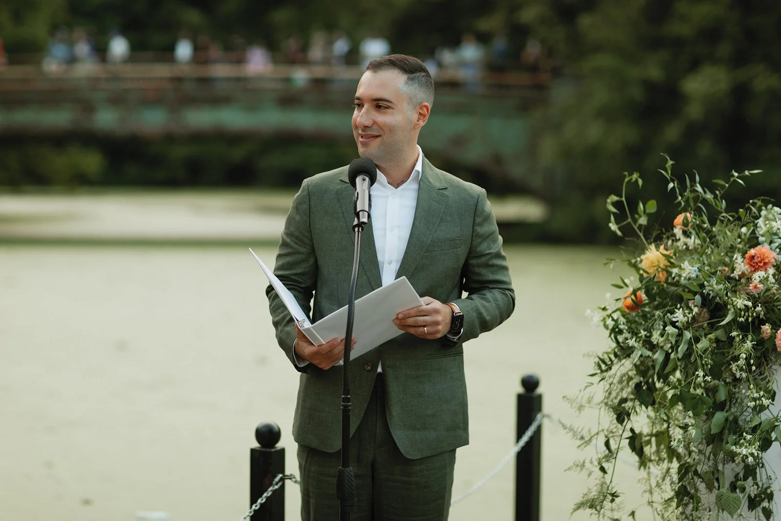 A man in a gray suit holding papers, speaking at a microphone during an outdoor event near a body of water with a bridge and a floral arrangement nearby.