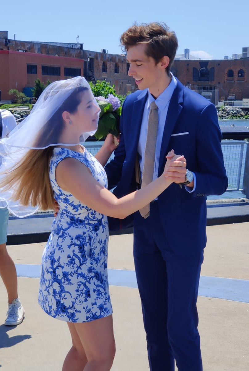 Young woman in a floral dress with a veil holds a bouquet of flowers, dancing with a young man in a blue suit outdoors.