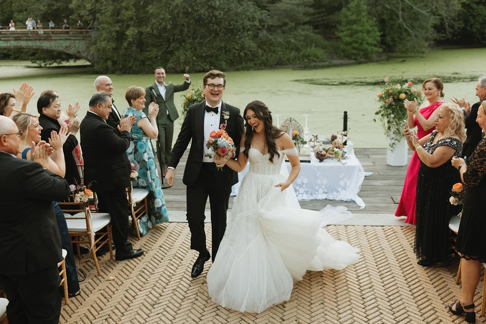A newlywed couple walking hand-in-hand on an outdoor wedding deck by a pond with green water, surrounded by friends and family applauding and smiling.