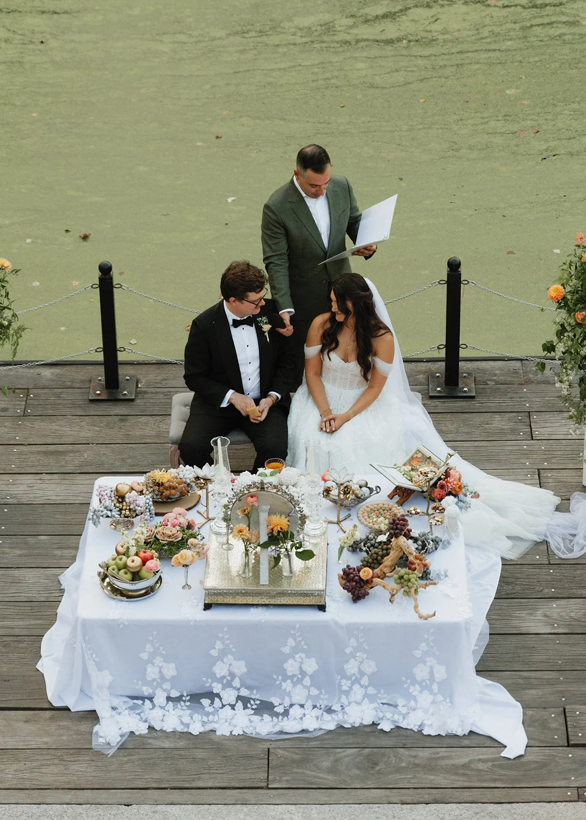 A wedding ceremony taking place outdoors on a wooden dock over a pond with green water. A bride in a white wedding dress and veil and a groom in a black tuxedo are seated at a decorated table with flowers, grapes, and other items. An officiant in a gray suit stands behind them holding a folder, celebrating the marriage.