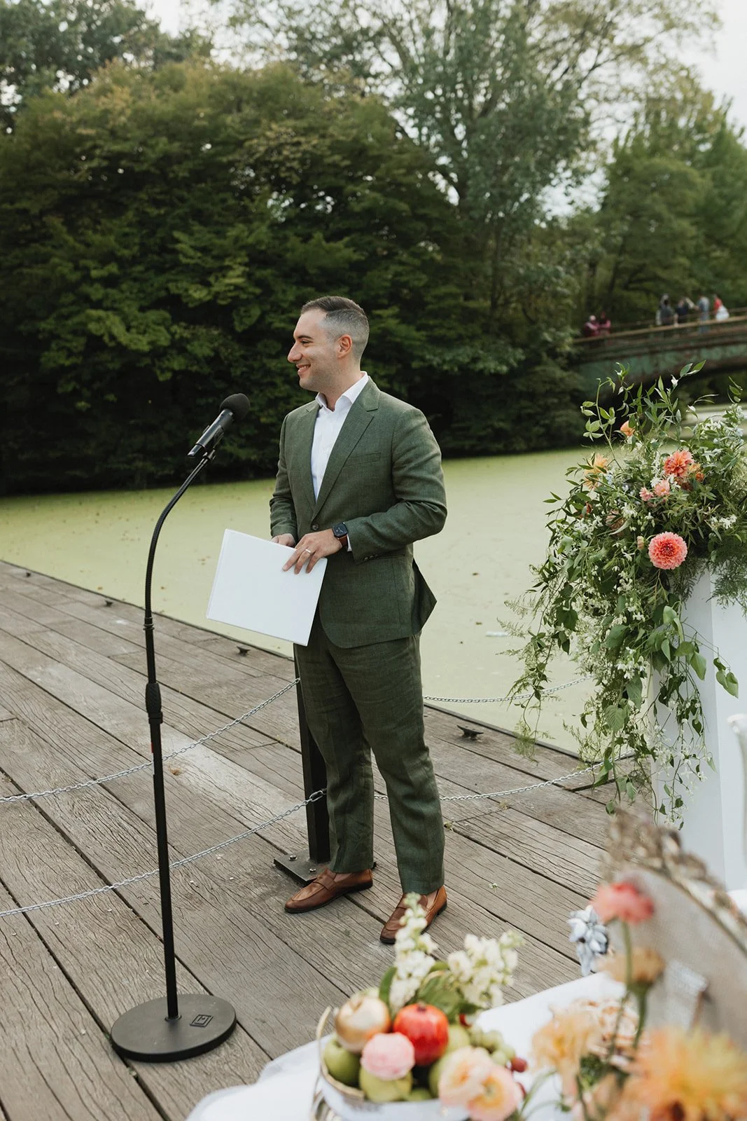 A man in a green suit giving a speech outdoors near a pond with green algae on the surface, surrounded by trees, with a floral arrangement and apples on a table nearby.