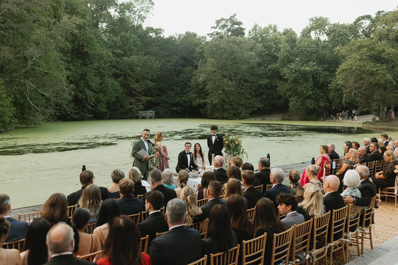 Wedding ceremony taking place outdoors on a lakeside dock, with guests seated and a officiant reading, surrounded by trees and water covered with green algae or duckweed.