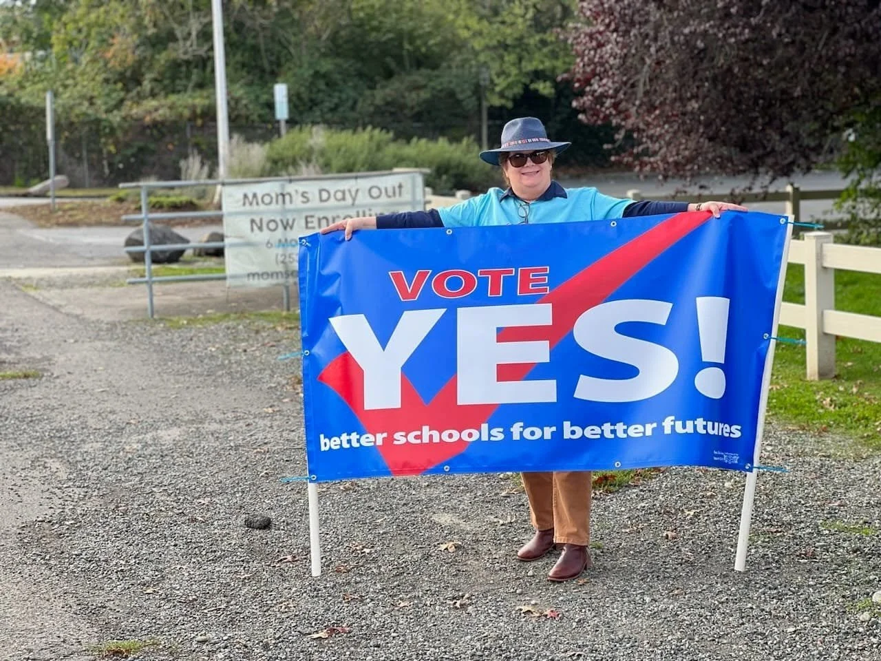 A woman holding a large blue campaign sign that says "VOTE YES! better schools for better futures" at an outdoor event. She is wearing sunglasses, a wide-brimmed hat, a blue shirt, and brown pants, with trees and a fence in the background.
