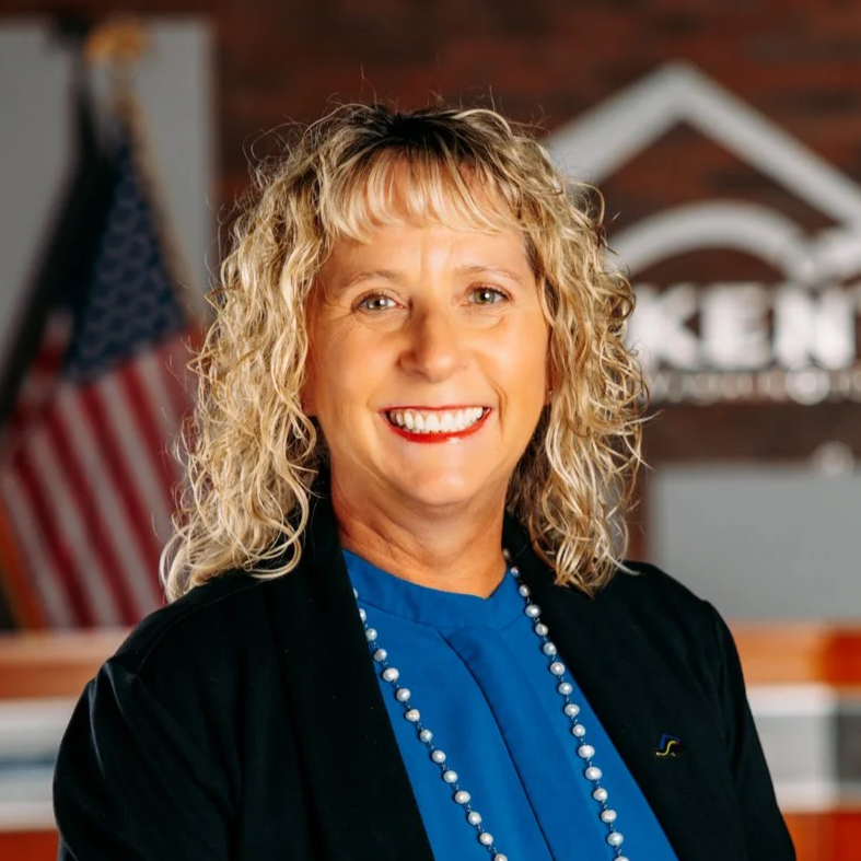A woman with blonde curly hair smiling, wearing a blue top, pearl necklace, and black blazer, standing in front of an American flag and a wooden background with a logo.
