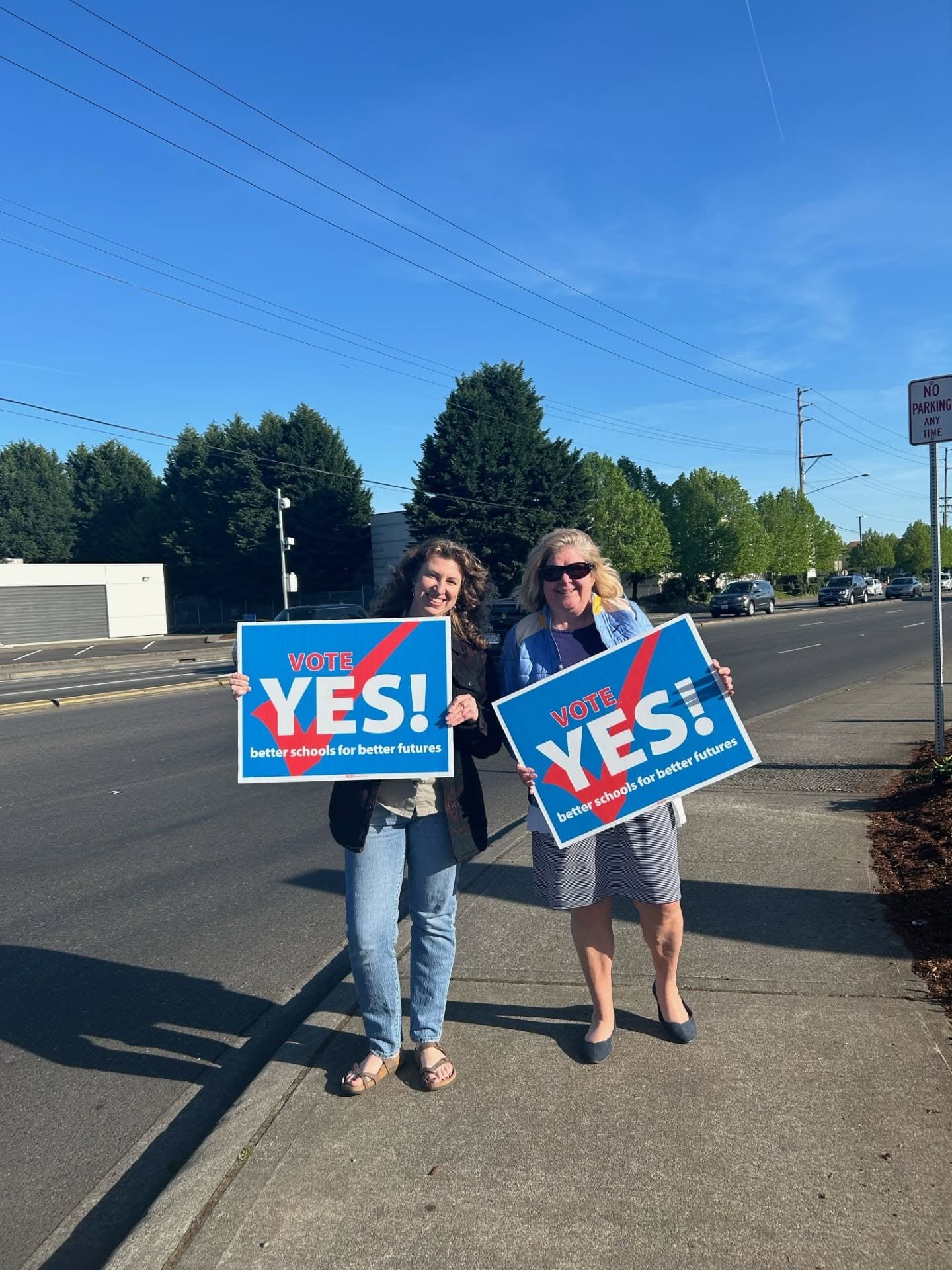 Two women standing on a sidewalk holding signs that say "Vote Yes! Better schools for better futures." They are smiling and appear to be participating in a campaign or protest supporting education.