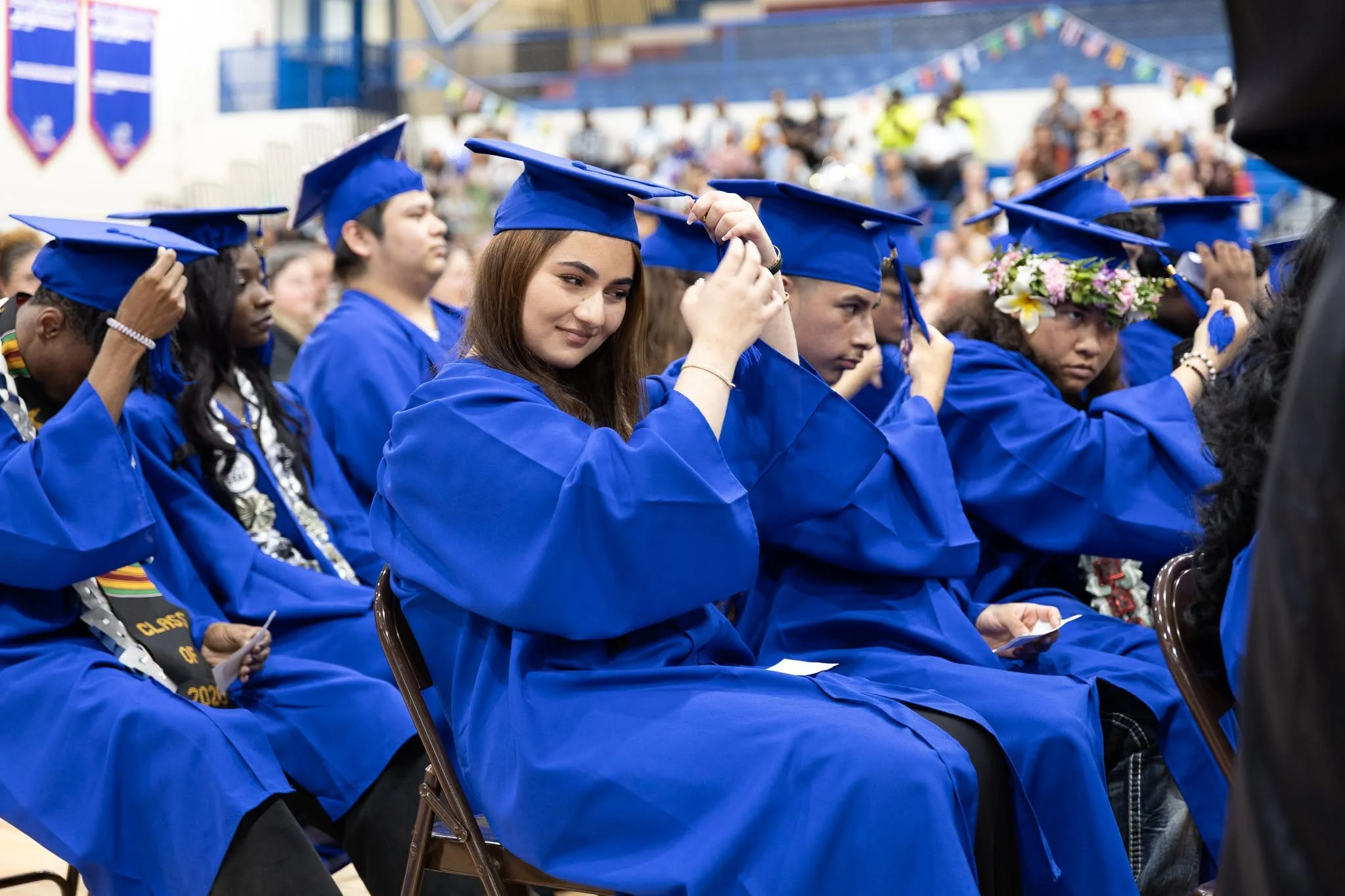 Group of graduates in blue caps and gowns sitting in chairs during a ceremony, with some adjusting their caps and a woman wearing a floral crown.
