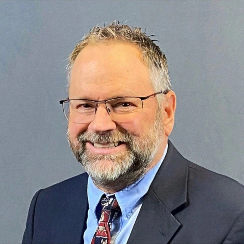 Portrait of a smiling middle-aged man with glasses, a beard, wearing a suit, blue shirt, and patterned tie against a gray background.