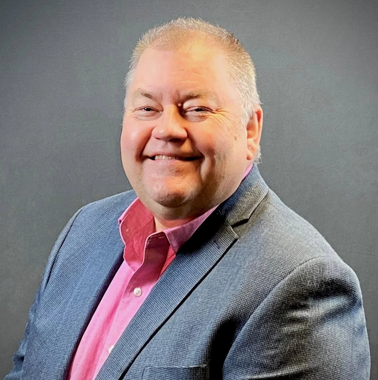 Portrait of a smiling man with short, graying hair, wearing a gray blazer and a pink collared shirt, against a dark gray background.